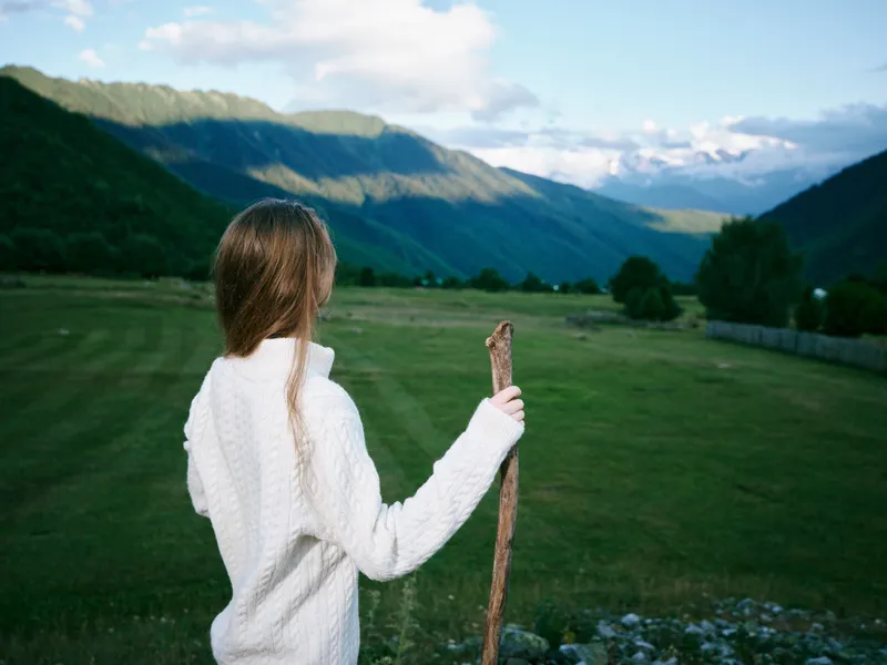 Girl in a white sweater stands in a green field, holding a wooden stick, with distant mountains and blue sky, capturing a quiet moment of outdoor exploration and open horizon