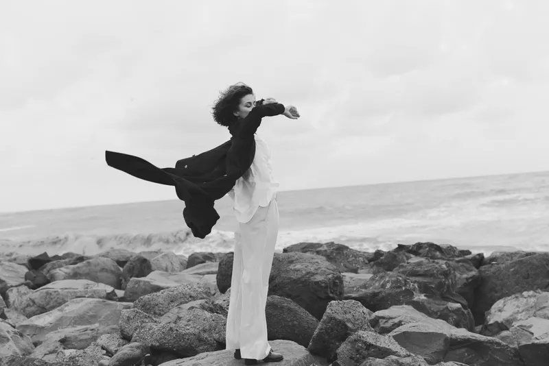 Windy dress flows as a figure stands on rocky coast by the sea, wind and waves create a serene monochrome scene with graceful movement