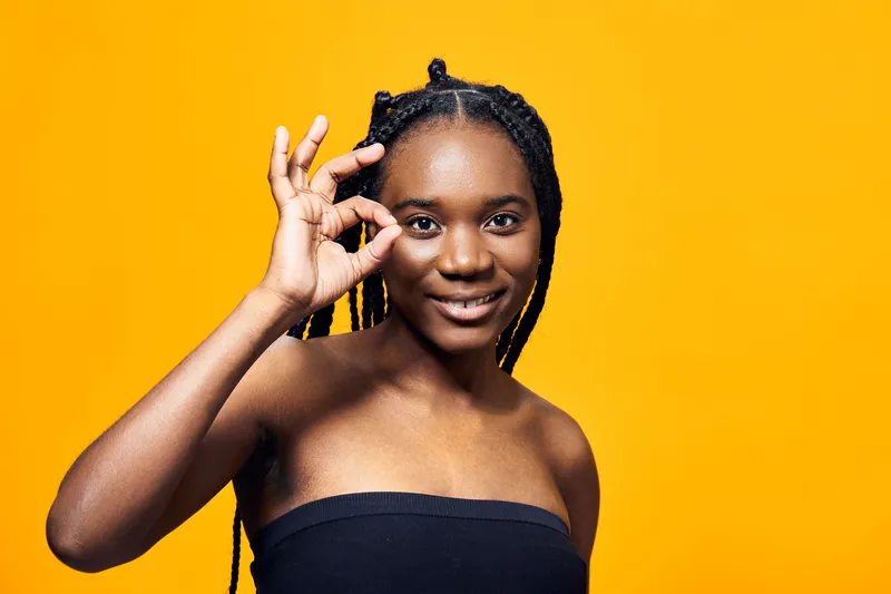 Young Black woman with braided hair giving an OK sign against a bright yellow background, exuding positivity, confidence, and joy in a vibrant setting