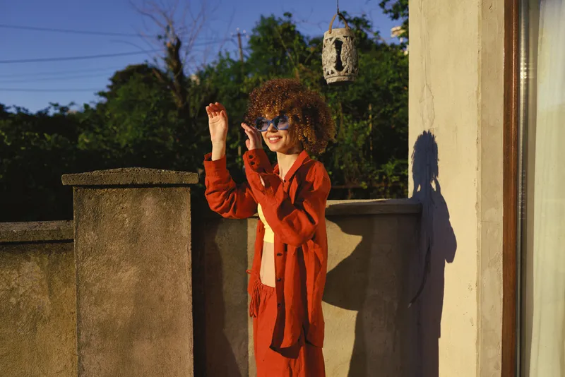 Woman wearing red suit enjoys a sunny outdoor moment on a terrace, lifestyle portrait with candid smile, cheerful mood, fashionable look, relaxed pose, natural light, shadow play, casual elegance