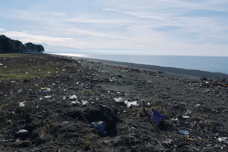 Pollution, beach, litter, garbage, environment, coastline, shoreline, waste on a rocky shore with scattered trash and a calm sea in the distance.