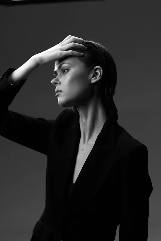 Black and white portrait of a thoughtful woman in a blazer with hand on forehead, showing emotion and elegance in studio lighting.