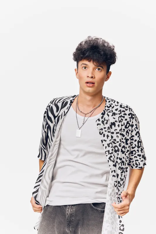 Young man wearing a patterned shirt looks surprised with open mouth and wide eyes, standing against a plain white background, casual style and expressive mood.