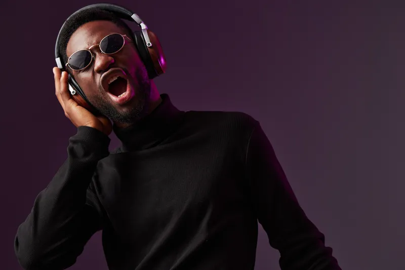 Young Black man singing passionately, wearing headphones and sunglasses, against a dark purple background, exuding emotions of joy and freedom