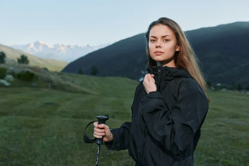 A young woman in a black jacket stands outdoors in a grassy valley with distant mountains. She holds a trekking pole and gazes at the camera with a calm, confident expression.