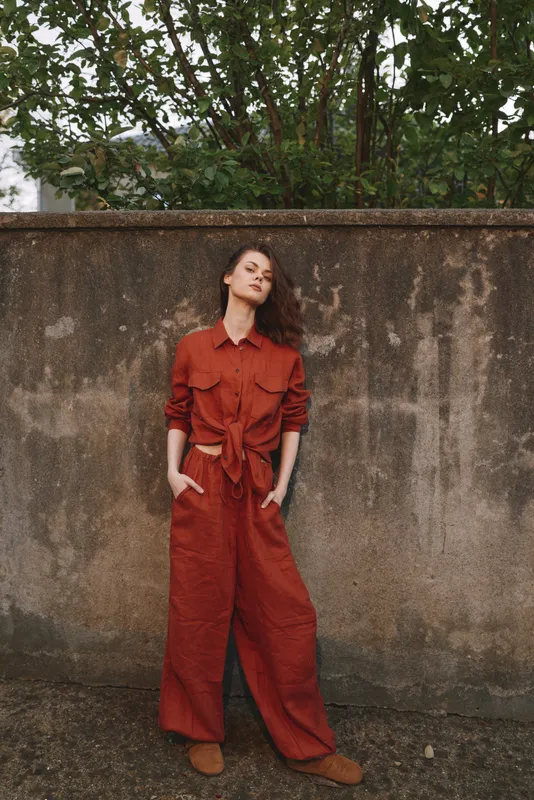 Vibrant woman outdoors in a rust jumpsuit, relaxed pose, subtle smile, natural light, soft breeze, urban backdrop, earthy tones, fashion forward lifestyle moment near a weathered wall