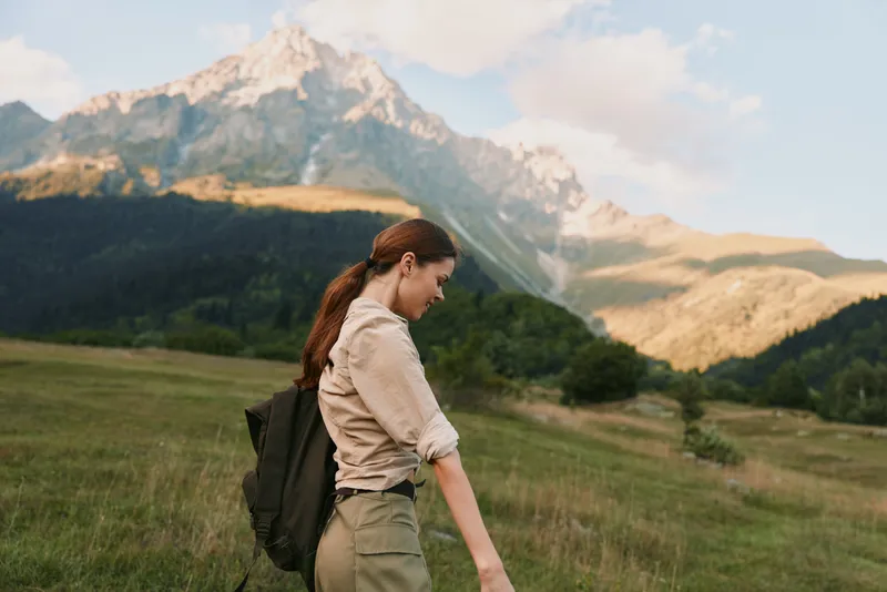 Hiker woman with backpack walks through a sunlit meadow toward distant mountains, enjoying a calm outdoor moment