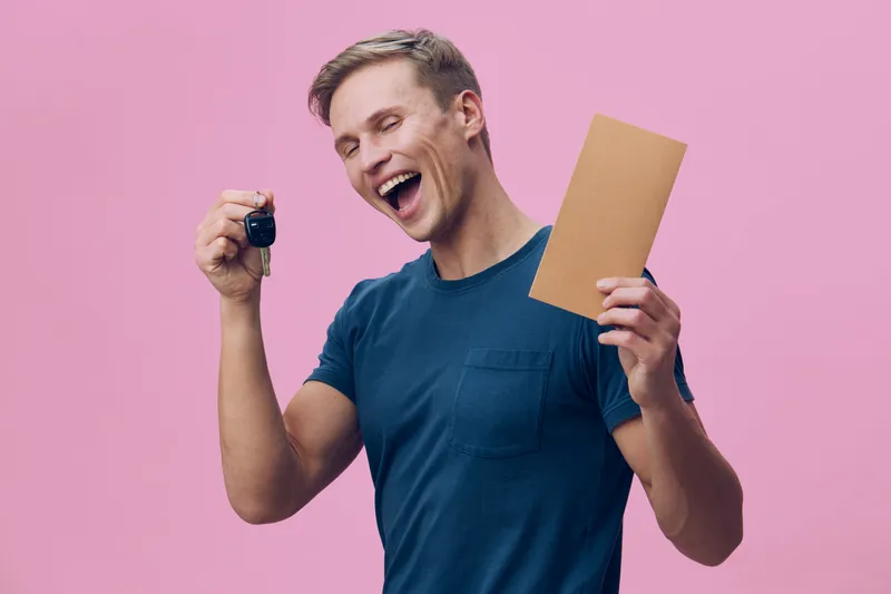 Excited happy smiling young man holding car key and brown envelope isolated on pink background joy emotion lifestyle concept