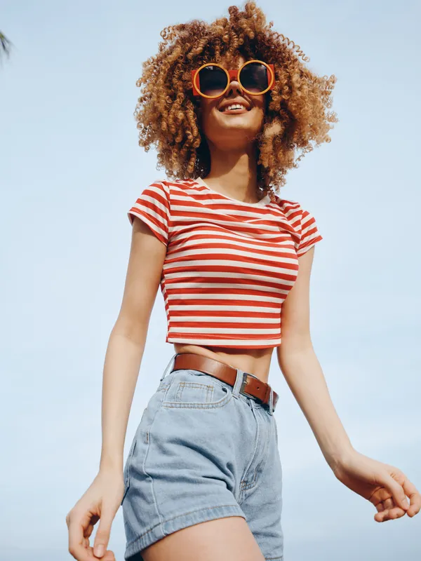 Young woman with curly hair wearing a red and white striped crop top, denim shorts, and oversized sunglasses stands against a bright blue sky, exuding carefree summer style for photography.