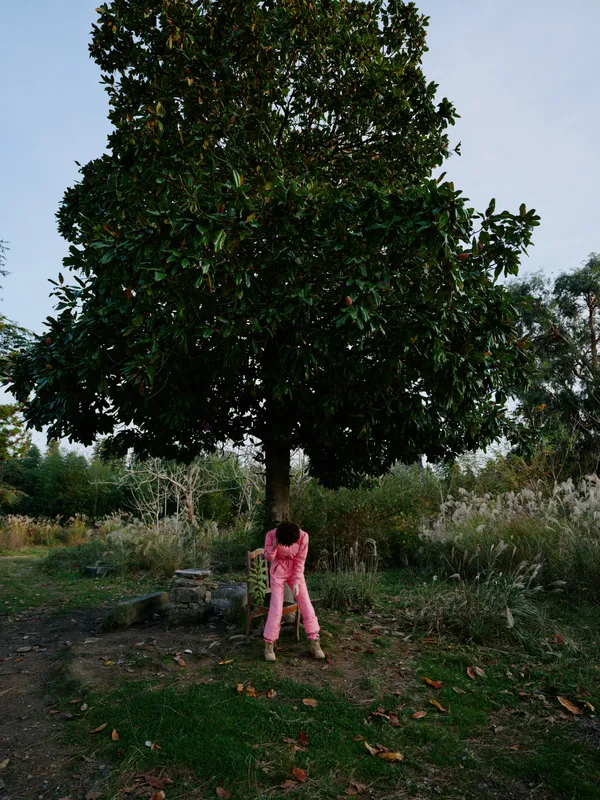 Child in pink clothing stands beneath a large tree in a park, autumn leaves scattered on the ground, conveying exploration and connection with nature.