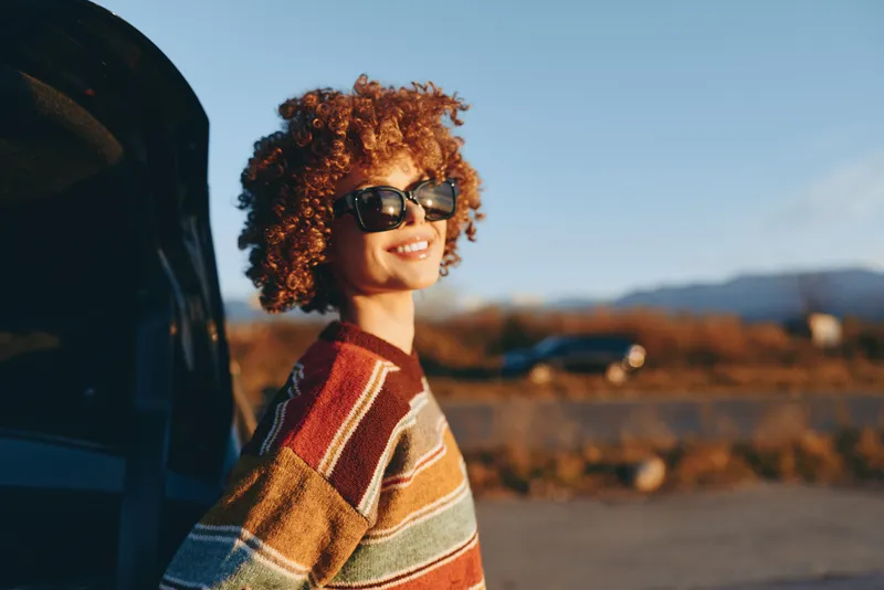 Woman lifestyle outdoors, curly hair, sunglasses, smiling beside car during roadtrip, striped sweater, warm autumn daylight, scenic countryside background, relaxed mood
