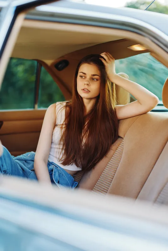 A young woman with long brown hair sitting in a vintage car, expressing a thoughtful mood, wearing a casual outfit, reflecting a summer vibe.