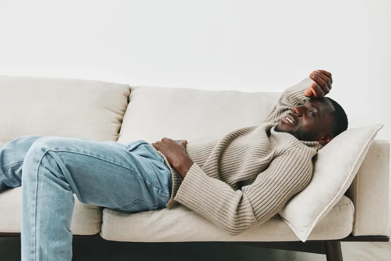 A young Black man in a cozy sweater lounging on a sofa, expressing relaxation and tranquility in a minimalist setting with soft colors
