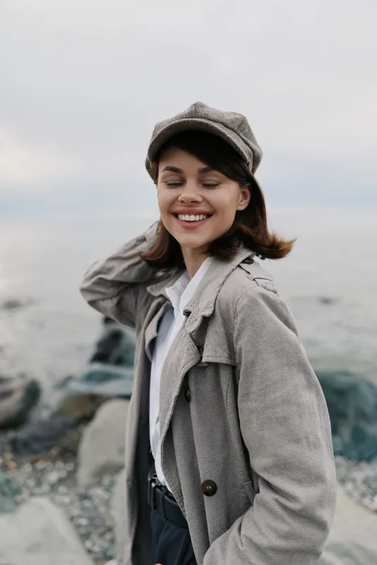 Smiling young woman in textured coat and hat stands by rocky shore with eyes closed. Casual autumn fashion and natural beauty captured in soft outdoor light by the sea.