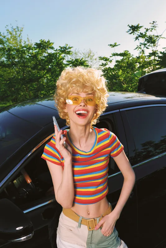 Cheerful woman with curly blond wig wears striped crop top beside a dark car, talking on a phone, retro lifestyle portrait with bright sunlight and playful mood and bold colors.