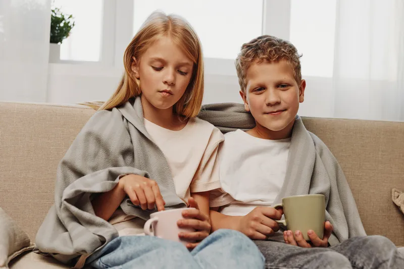 Two children wrapped in a blanket sit on a sofa drinking hot beverages. The cozy indoor scene shows a calm boy and girl enjoying warmth and comfort at home.