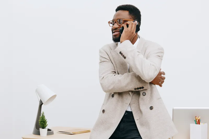 Professional young African American man with glasses wearing beige blazer talking on smartphone while standing at desk with laptop and plant in modern office space isolated on light background. Busine