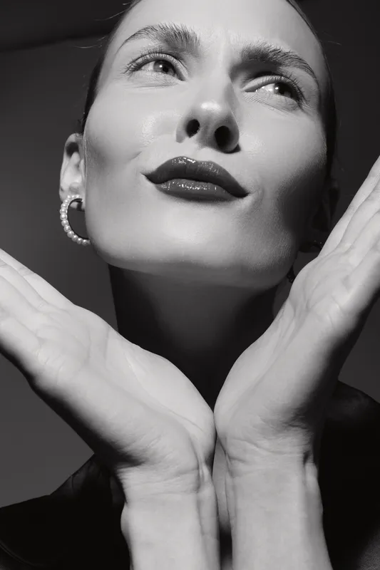 Close-up portrait of a fair skinned Caucasian woman with slicked hair and pearl earrings, posing with hands near face in dramatic black and white studio lighting. Concept of elegance and beauty.