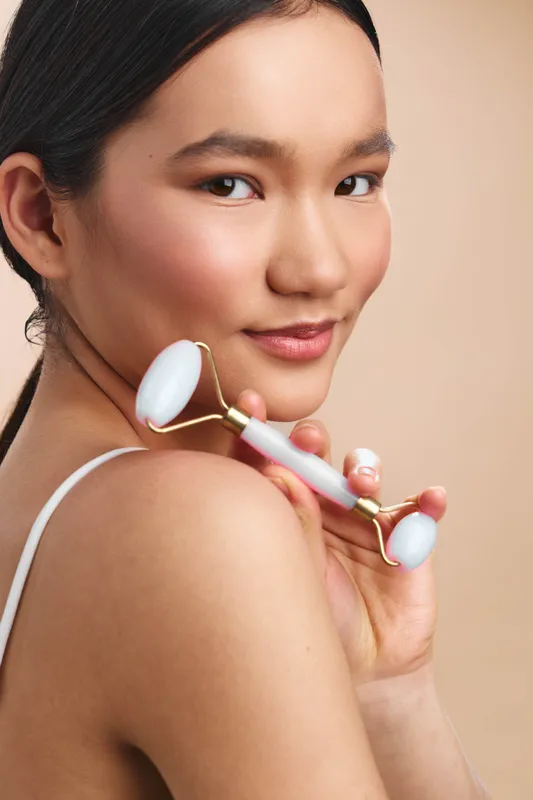 Asian woman uses a facial roller in her skincare routine. She smiles softly, skin glowing and relaxed, captured in a warm, minimalist setting, highlighting selfcare and a calm beauty moment.