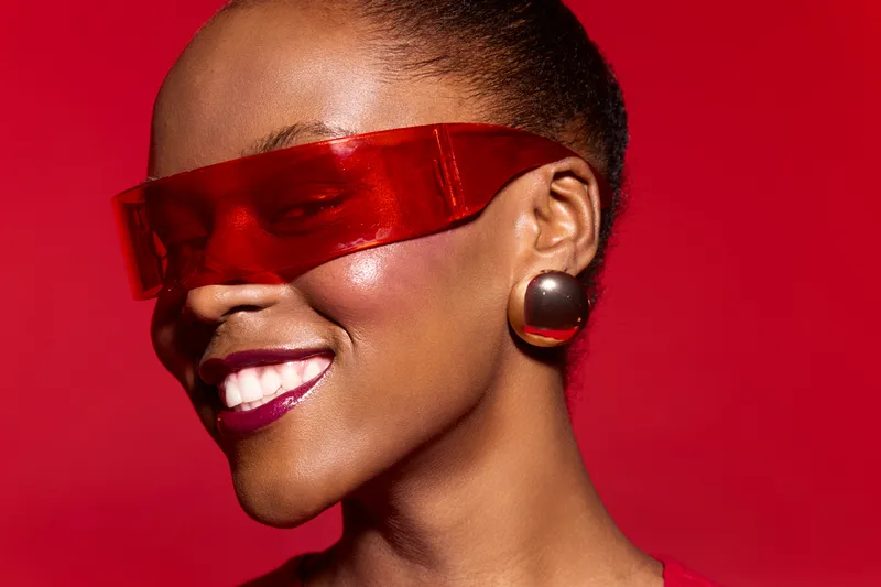 Close-up of a smiling African woman with dark skin wearing futuristic red glasses and large earrings. Bright red background enhances the vibrant and confident mood of the portrait.