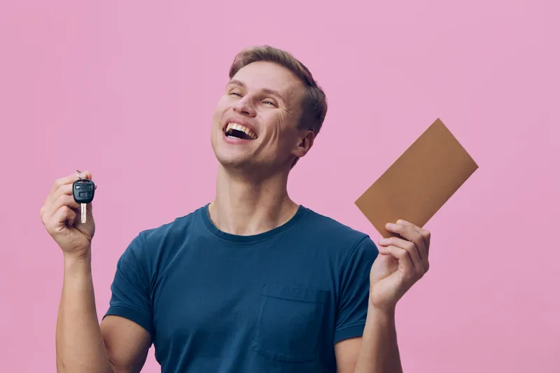 Smiling happy young man wearing casual blue t-shirt holding car key and passport with brown cover, celebrating success against pink background. People lifestyle concept, travel, achievement, joy, exci