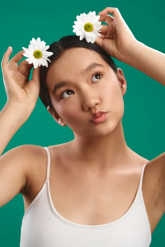 Asian woman in a white tank top playfully arranges daisies in her hair against a vibrant green studio backdrop, delivering a cheerful, fresh portrait that radiates creativity and natural charm.