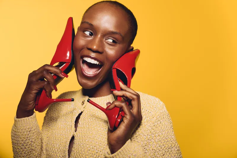 Happy dark skinned African woman holds red high heels near face, smiling and looking away. Bright yellow background, casual sweater, concept of fashion, joy, and beauty.