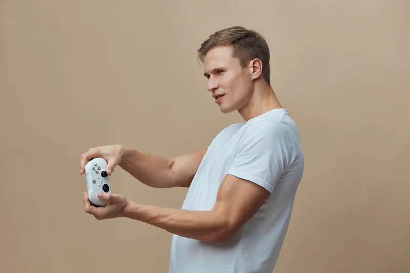 Man holding game controller, playing video games, casual look, light blue t-shirt, concentrated expression, indoor studio shot, beige background, leisure activity, entertainment concept.