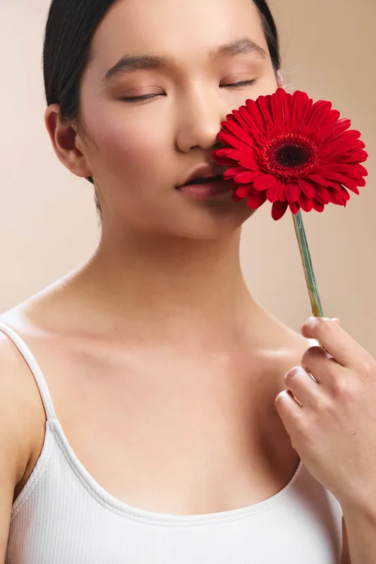 Asian woman closes eyes as she holds a vibrant red gerbera near her face, creating a serene close-up portrait with soft lighting and minimalist styling.