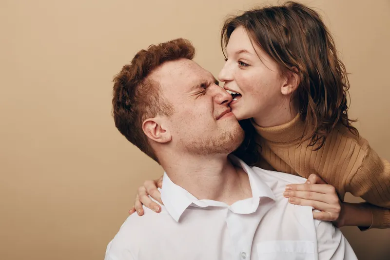 Happy young couple in casual clothes share playful moment, woman hugging man from behind on beige background. Concept of love, relationship, joy, and intimacy between partners.