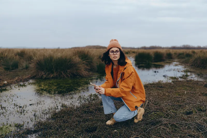 Woman lifestyle in nature, sitting on dry grass by a pond, wearing orange jacket and knit hat, looking at camera, calm morning in autumn landscape, candid travel portrait, with reeds behind