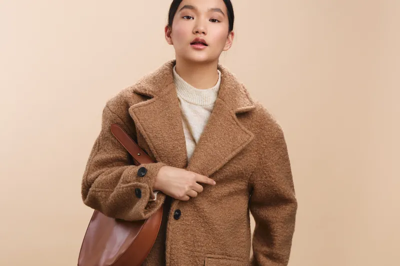 asian woman wearing a cozy coat stands in a neutral setting, presenting a calm, stylish vibe. she looks toward the camera, exuding confidence and approachable professionalism.