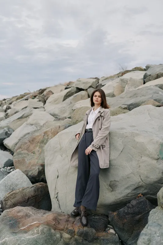 Young woman in grey coat and dark pants stands on rocky shore under cloudy sky. Casual fashion and outdoor lifestyle concept with natural landscape and relaxed mood by the sea.