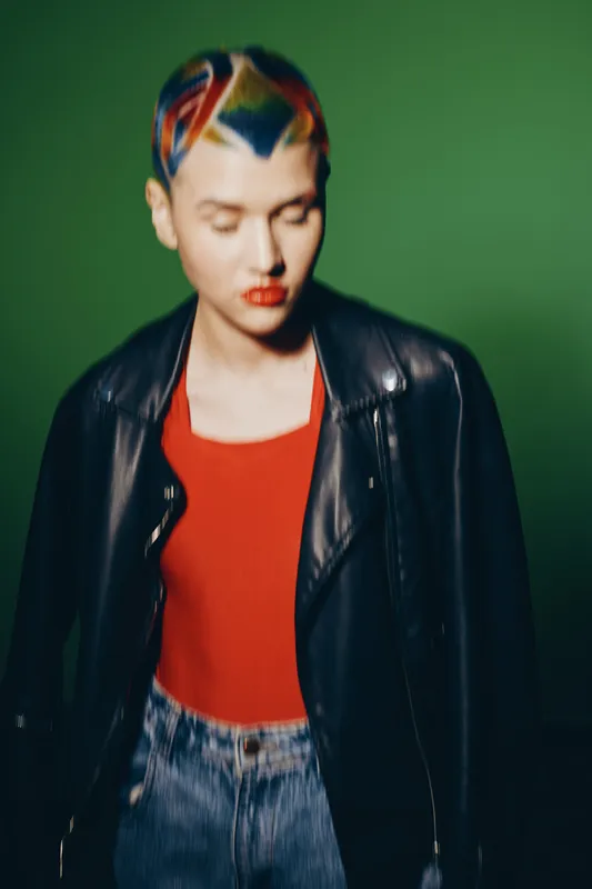 woman with colorful short hair wearing a black leather jacket and red top posing against a green background in casual style
