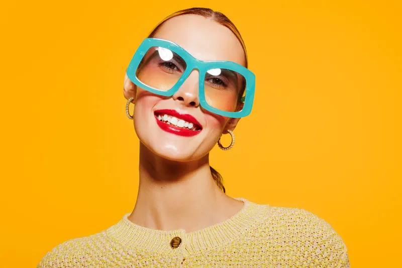 Smiling woman with bright red lipstick wears large blue sunglasses and yellow sweater against vibrant orange background. Concept of fashion, summer style, and confident beauty.