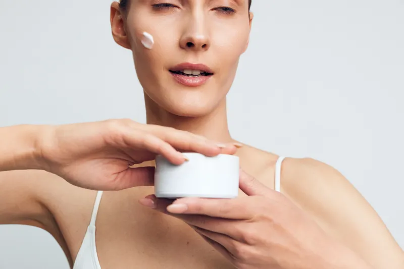 Young woman with short hair applies face cream on her cheek while holding a white cosmetic jar against a plain light background in a skincare routine.