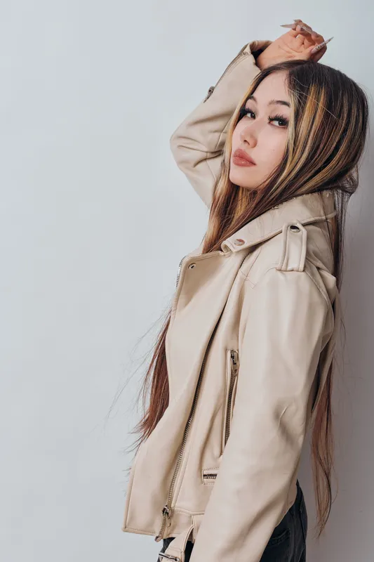 Young woman with long hair and beige leather jacket posing in profile with hand raised, looking confidently at camera against a plain light background in studio.