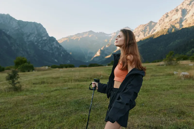 A woman hikes in a mountain landscape, standing in a grassy valley with a trekking pole, wearing a jacket and crop top. She gazes toward distant peaks, embodying adventure and outdoor resilience.