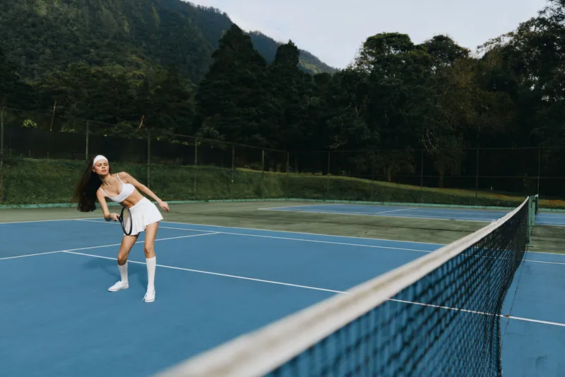 Young woman playing tennis on a blue court surrounded by lush greenery, showcasing determination and joy in her athletic wear