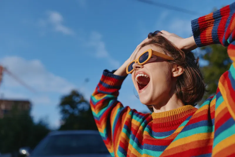 Woman in striped sweater and yellow sunglasses outdoors, young adult smiling and joyful, hands in hair, colorful casual outfit, urban sunny day travel lifestyle portrait