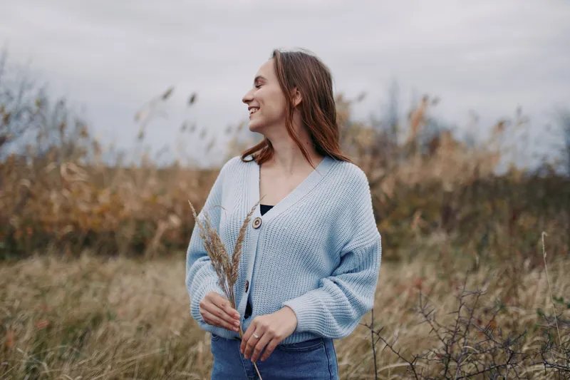 An authentic portrait of a smiling woman standing in a natural field, wearing a light blue cardigan, holding tall grasses, and appearing calm, confident, and genuine.