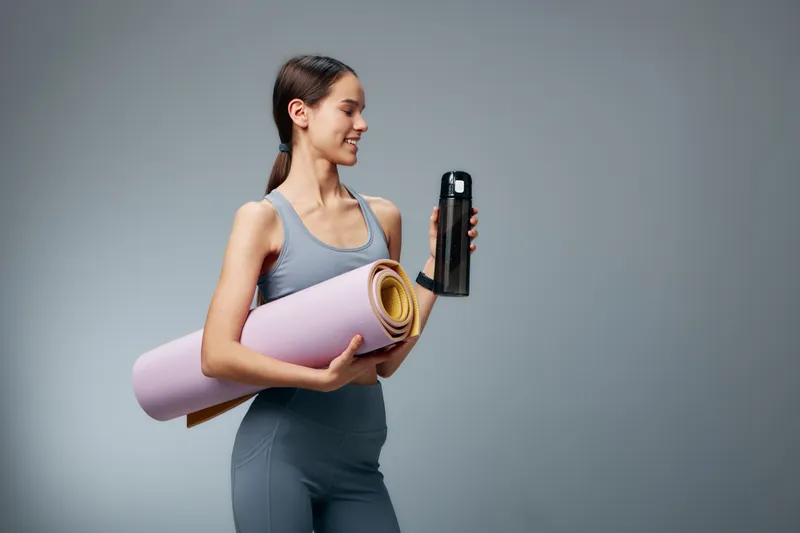 Fitness woman in athletic wear holding a yoga mat and water bottle, ready for a workout. Clean studio backdrop highlights health, active lifestyle, and dedication to training and well being.