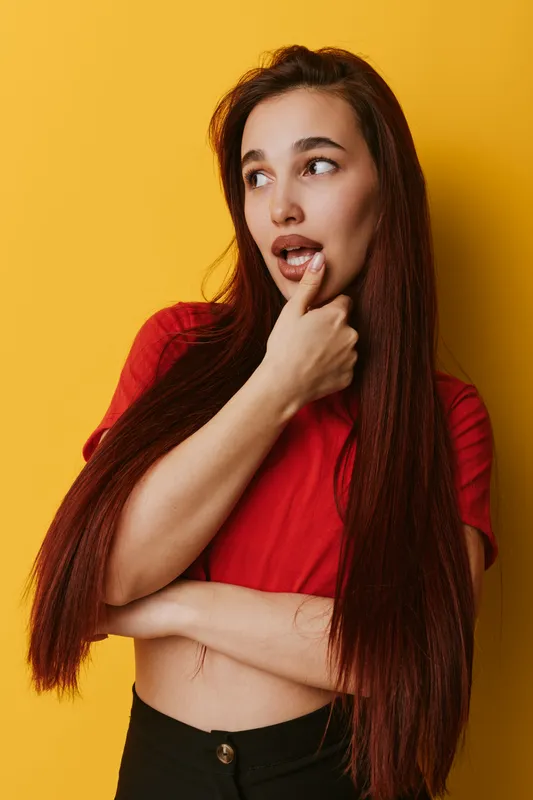 Young woman with long hair wearing red shirt standing against yellow background looking sideways with thoughtful expression and finger on chin in casual pose for contemplation or decision.