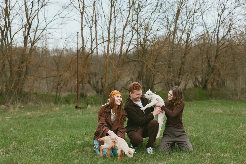 Smiling family outdoors in a grassy field with a playful goat kid, sharing a lighthearted moment. Casual clothing, natural light, modern lifestyle scene focused on joy, animals, and connection,