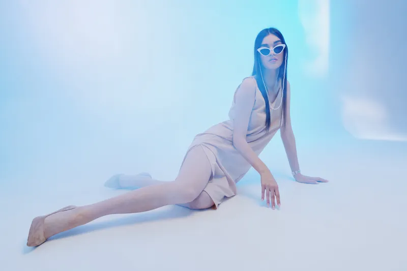 Fashionable woman in a beige dress and white sunglasses poses on the floor in a bright studio with blue lighting. Concept of beauty, style, fashion, and modern elegance.
