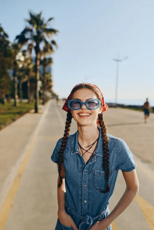 smiling woman in blue denim dress with braided hair and large sunglasses enjoys sunny day outdoors near palm trees