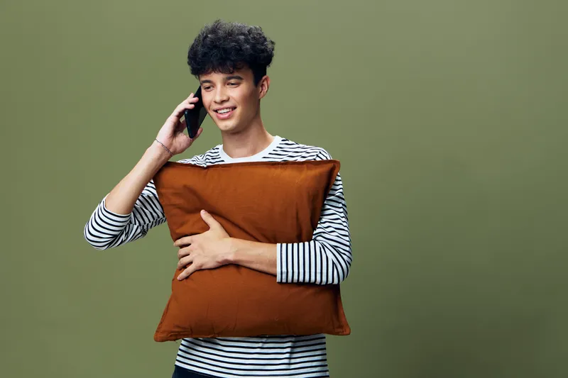 smiling man talking on phone holding brown pillow with striped shirt on olive green background, casual lifestyle moment