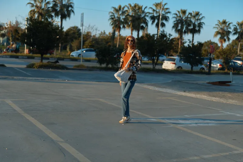 Woman in parking by palms with sunglasses and bag, casual street outfit in warm golden hour glow; candid authenticity and mindful living vibe capturing emotional storytelling of modern city life.