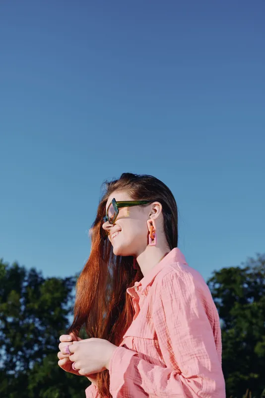 Young woman enjoying a sunny day outdoors, wearing stylish sunglasses and a pink shirt, with long hair flowing in the breeze under a clear blue sky
