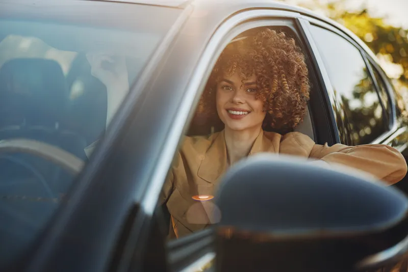 Bright-eyed woman with curly hair sits in a car, smiling toward the camera. Natural light spills through the window, creating a warm retro mood and casual lifestyle.